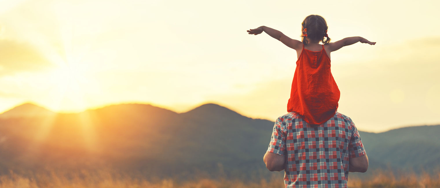 A man gives his daughter a shoulder ride looking out over a mountain sunset