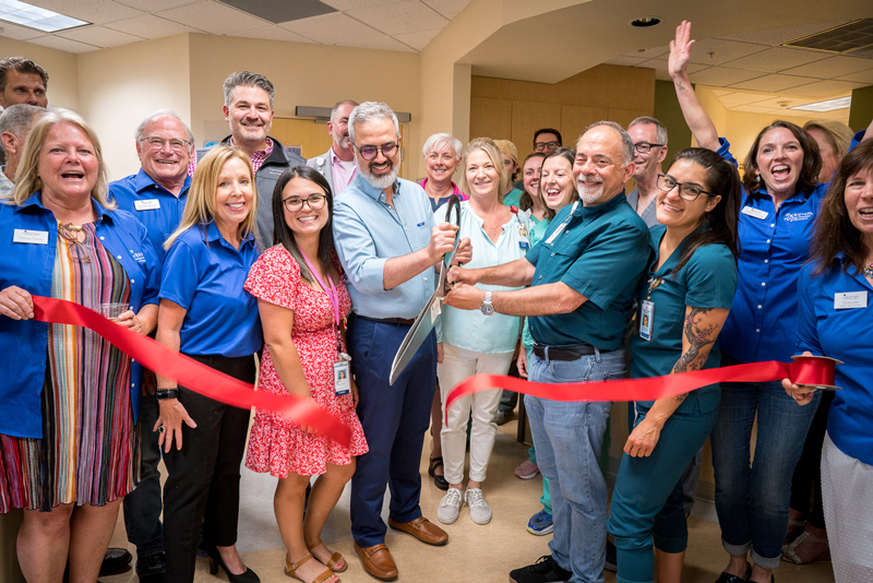 Two men slice through a big red ribbon as a group of doctors and nurses cheer and clap