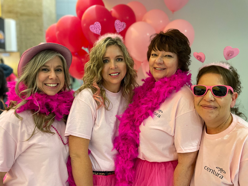 Four women dressed in pink t shirts, feather boas, and hats pose in front of balloons in shades of pink and red
